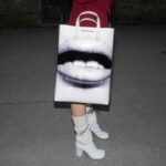 Close-up of a stylish bag with art print, paired with high heels on a nighttime pavement.