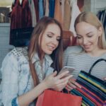 Two women enjoying a shopping spree and sharing a moment on their smartphone in a clothing store.