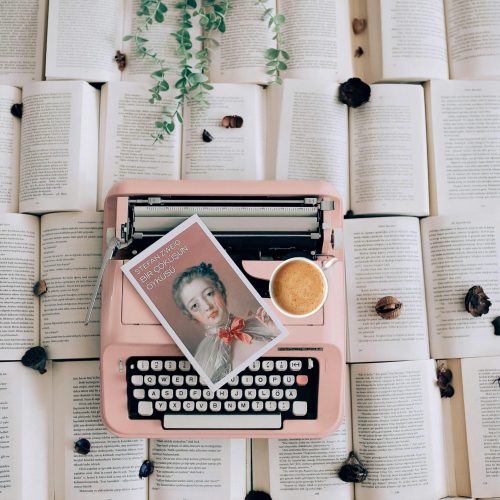 A pink typewriter on open books with a coffee cup and plant, offering a vintage literary vibe.